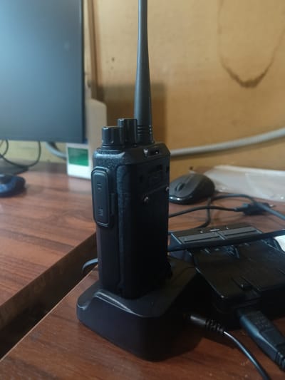 A black two-way radio is sitting in its charging dock on a wooden desk. The radio is upright, with its antenna extending upwards. The charging dock is black and has several slots, with cables plugged into it. To the left of the radio, a portion of a computer monitor is visible. In the background, a wall with some staining can be seen. A computer mouse is also present on the desk, to the right of the radio. The lighting suggests it is daytime, and the scene appears to be indoors. There are no people visible in the image. There is no visible text in the image.