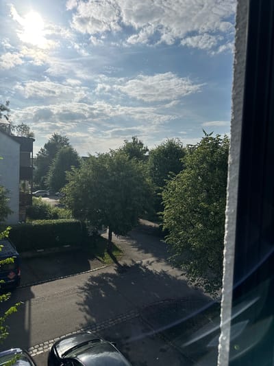 This is an outdoor, eye-level, medium shot of a residential street in Rosenheim, Germany. The image is dominated by trees and the sky, with a partial view of a building and parked cars. The sky is partly cloudy with bright sunlight filtering through. The trees are lush and green, casting shadows on the street. A dark-colored van and two cars are parked along the street. The building on the left appears to be an apartment complex with balconies. The scene suggests a calm, suburban environment on a sunny day.