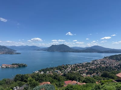 The image captures a scenic vista of Lake Maggiore in northern Italy, likely taken from an elevated viewpoint above the town of Someraro. The sky is a clear, vibrant blue with scattered white clouds. The lake dominates the center of the frame, reflecting the sunlight. In the foreground, lush green trees and the terracotta rooftops of houses are visible, suggesting a residential area. The midground features a densely populated town with buildings clustered together, nestled along the shoreline. A notable feature in the lake is a small, picturesque island with buildings and manicured gardens. In the background, rolling hills and mountains form a majestic backdrop, their slopes covered in vegetation. The overall impression is one of a tranquil and beautiful lakeside landscape on a sunny day.