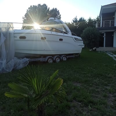 In the city of Limani Litochorou, Greece, a white motorboat is parked on a trailer in a grassy yard, situated in front of a modern, dark-colored house with a white railing on its upper balcony. The sun is setting behind a line of trees, casting a strong glare that obscures the upper portion of the boat. The boat is covered with a transparent tarp on its left side. The yard is covered in green grass with a small palm-like plant in the foreground. The trailer has three sets of wheels.