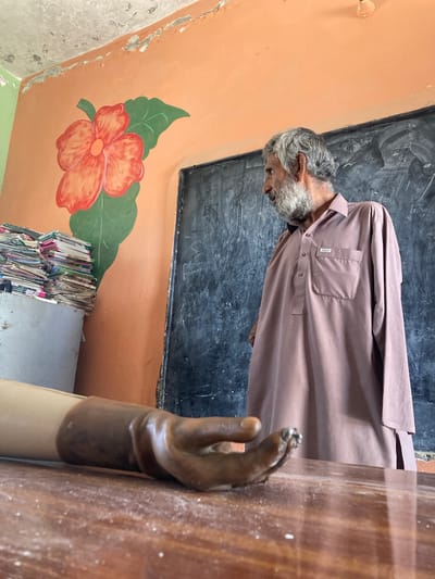 The image shows a man with gray hair and a beard, wearing a long brown shirt, standing in a room with an orange wall, a blackboard, and a painted red flower. The room appears to be in a state of disrepair. In the foreground is a prosthetic arm on a wooden table. The location is Qilla Saifullah, Pakistan.