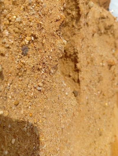 The image displays a close-up view of loose, coarse sand with visible grains of varying sizes and colors, predominantly shades of brown and tan, interspersed with some lighter-colored pebbles and darker, possibly mineral, flecks. The sand appears to be dry and somewhat clumpy, forming uneven textures and small crevices. The lighting suggests it is outdoors, casting subtle shadows. The scene is devoid of any human presence or identifiable landmarks. This image captures a texture-rich detail of the natural ground surface in Kuru, Nigeria.