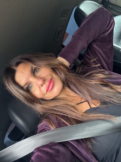 A woman with long, wavy brown hair is positioned inside a car, leaning her head back against the headrest. She is wearing a dark purple velour tracksuit and a black tank top with a thin gold necklace. A grey seatbelt is fastened across her chest. Her eyes are open, looking towards the camera, and she has bright pink lipstick on. The background shows the car's interior, including a black leather seat and part of a headrest. The lighting suggests it is daytime, and the image appears to be a selfie taken in the car. The scene does not contain any elements that suggest a specific location within the city of Кварса, Russia.