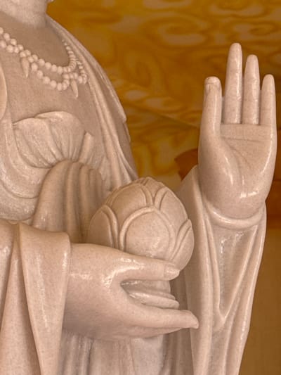 The image displays a close-up view of a light-colored stone statue, likely marble, located in Đăk Mar, Vietnam. The statue's upper torso and arms are depicted, featuring detailed drapery of robes. A double-stranded necklace with bead-like elements and pointed pendants is visible on the left side of the figure's chest. The figure's right hand is raised to shoulder height, fingers extended upwards with the palm facing forward. The left hand holds a closed lotus flower, cupped by the fingers. The background is a blurred, warm yellow-orange with subtle, indistinct patterns.