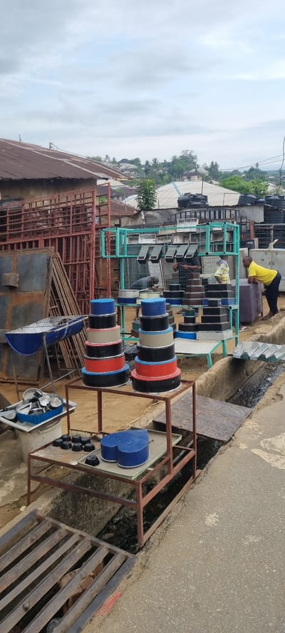 This media file depicts an outdoor metal fabrication or sales workshop, likely located in Calabar, Nigeria, during the daytime under an overcast sky.
The scene shows several individuals engaged in activities related to metalwork. In the background, a man in a yellow t-shirt and dark trousers is leaning over a large, black cylindrical object, possibly a tank. Near the center, another man wearing a plaid shirt is focused on arranging or working with smaller metal items on a display stand.
The workshop is filled with various metal products. Large black water tanks are visible in the background on the left. In the mid-ground, numerous light-colored metal sheets, possibly roofing or paneling, are neatly stacked on green metal racks. Dominating the foreground and center are several sturdy metal stands displaying colorful, circular metal objects in blue, black, red, beige, and silver, which could be industrial spools, decorative components, or covers. Below these, smaller blue heart-shaped or shield-shaped metal pieces and small black circular knobs or caps are arranged on separate display structures. A blue metal structure in the lower left foreground has the visible text "GE. ELECTRICAL". Various reddish-brown metal frames, rebar, and construction materials are also scattered around the site.
The workshop is set beside a paved road or path, with a drainage ditch covered by a metal grate running along its edge. The background features green tropical vegetation and residential buildings, all under a bright but cloudy sky. The overall impression is that of a bustling, functional outdoor commercial or industrial space.
