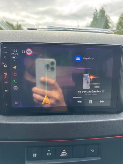 This is a close-up, rotated shot of a car's infotainment screen. The screen displays music playback, with album art of a wolf and text in Russian. The song title is "не расслабляйся" (don't relax), and the artist is "Скриптонит". There are playback controls visible: pause, next track, and previous track.

Above the music interface, there's a section labeled "На работу" (to work), accompanied by a blue briefcase icon. At the bottom of the screen, a status bar shows the time as 11:36, LTE connectivity, and a battery indicator. To the left of the music, speed indicators show 74 and 90, with 90 inside a red circle, possibly indicating a speed limit or alert.

The reflection on the screen shows a person's hand holding a smartphone, with its camera lenses clearly visible. The background outside the car, seen through the windshield, suggests a daytime scene with bright, overcast skies and some greenery, hinting at a temperate climate. The interior of the car, with textured plastic panels and red trim, is also visible. The overall impression is a snapshot of someone interacting with their car's media system while possibly on the go, given the "to work" label and speed indicators.

Visible text includes: "MIC", "RST", "11:36", "LTE", "74", "90", "На работу", "не расслабляйся", "PARENTAL ADVISORY EXPLICIT CONTENT", and "Скриптонит". The context of Parma, Russia is provided separately.