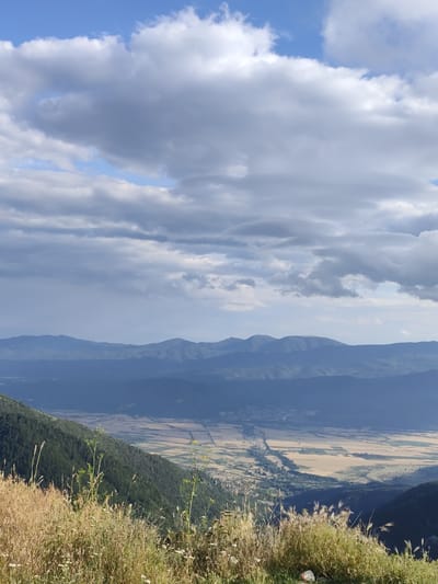 This media file captures a scenic vista from a high vantage point, overlooking a valley and distant mountains. The foreground is dominated by dry, golden grasses and wildflowers, suggesting a warm, dry climate. The middle ground reveals a lush green mountainside covered in dense trees, with a clear division between the dense forest and the cultivated fields of the valley below. The valley itself is a patchwork of golden and light brown fields, hinting at agricultural activity. A small settlement is visible nestled within the valley, with roads and buildings. The background features layered mountain ranges, fading into a hazy blue silhouette against the sky.
The sky above is a dynamic canvas of blue interspersed with dramatic, heavy clouds, some appearing grey and dense, possibly indicating approaching weather or recent rain. The lighting suggests it is either late afternoon or early morning, with the sun casting shadows and highlighting the textures of the landscape.
The scene evokes a sense of vastness and natural beauty. The time of day, coupled with the cloudy sky, creates a mood of gentle drama and tranquility. There is no visible text in the image. The location is Hristo Danovo, Bulgaria, which is consistent with the mountainous terrain and agricultural valley depicted. The image appears to be a still photograph.