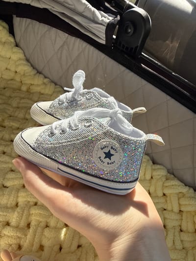 A pair of sparkly, silver baby sneakers are held in a person's hand. The sneakers have white laces and a white rubber toe cap. A circular patch with a blue star and "Baby" repeated around it is visible on the side of each shoe. The person's hand is partially illuminated by sunlight, casting a shadow on their skin.

The sneakers are positioned in front of a background that appears to be a light yellow, textured blanket. To the left of the sneakers, a quilted, grey material is visible, possibly part of a stroller or bassinet. The overall lighting suggests it is daytime, with natural sunlight creating bright highlights and soft shadows. The presence of the baby shoes implies a context related to infants or young children. The image does not provide specific details about the weather or a precise location, beyond the general context of Petropavlovsk-Kamchatskiy, Russia. There is no discernible action or event taking place beyond the presentation of the shoes.