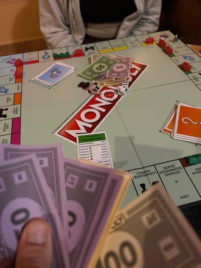The scene depicts an active game of Monopoly in an indoor setting within the Gerakas Municipal Unit, Greece. A light green Monopoly board, a Greek edition, is spread across a table. In the foreground, a person's hand, seen from a first-person perspective, holds a fan of Monopoly money, including purple M20 and M50 bills, and a yellow M100 bill. Across the table, another individual is partially visible from the chest up, wearing a light grey hooded sweatshirt over a dark top.
On the board, various game components are present:
* Red Monopoly houses are placed on multiple properties, notably three on a purple property on the left side and several others along the upper right edge. Green hotels are also visible.
* Stacks of game money, including purple M5, green M20, and yellow M50 denominations, are centrally located near the "MONOPOLY" logo.
* Two white dice displaying a '1' and a '2' (summing to 3) rest near the "MONOPOLY" logo, which features Mr. Monopoly.
* A stack of blue "Chance" cards (bearing a question mark shield) is positioned on the left side of the board.
* A stack of orange "Community Chest" cards (with a simple question mark) is on the right.
* A green "ΟΔΟΣ ΑΚΑΔΗΜΙΑΣ" (Academy Street) property card, displaying rent values from M26 to M1975, is positioned in the lower-middle section of the board.
* Visible board spaces include "ΦΟΡΟΣ ΕΙΣΟΔΗΜΑΤΟΣ ΠΛΗΡΩΣΕ" (Pay Income Tax) and "ΣΤΑΘΜΟΣ ΠΕΛΟΠΟΝΝΗΣΟΥ" (Peloponnese Station).
The environment suggests a casual gathering, with a blurred warm-toned wall in the background. The game is in progress, indicated by the arrangement of pieces and money.