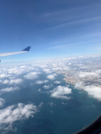 The image depicts a view from an airplane window, showing a portion of the wing, the sky, the sea, and a coastline with a densely populated city. The city appears to be situated along a beach. There is no indication of the location being Paiania Municipal Unit, Greece.