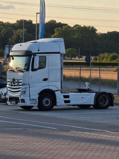 A white Mercedes-Benz Actros tractor unit is parked in a paved lot with visible lane markings. The truck has a license plate reading "DL-FS 915". In the background, a metal fence runs horizontally, separating the lot from a grassy area and a line of trees. The sky is a light orange-yellow, suggesting either sunrise or sunset, with power lines visible overhead. A blue flag on a pole is present behind the truck. Another car with license plate "KR-X 220" is partially visible to the left of the truck. The scene appears to be in an industrial or commercial area.