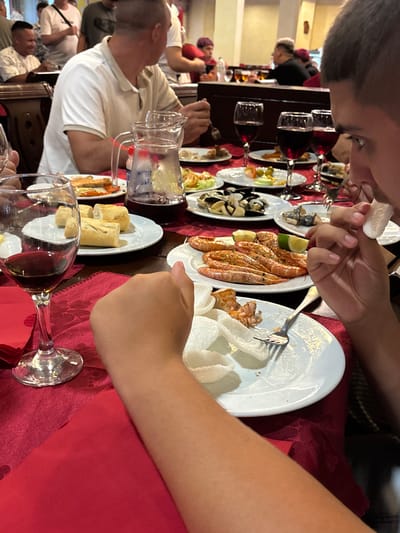 The image depicts a group of people dining at a restaurant, possibly in Molina de Segura, Spain, given the context. The scene is set at a table covered with a red tablecloth, laden with various dishes of food and drinks. In the foreground, a person's arm and hand are visible, holding a piece of white food, possibly shrimp cracker, towards their mouth. A glass of red wine and a plate with shrimp and lime wedges are also in the foreground. In the mid-ground, other diners are partially visible, some engaged in conversation, others eating. A pitcher of water and several plates with different food items, including bread, salad, and seafood, are spread across the table. The restaurant appears to be moderately crowded, with other patrons and staff visible in the background, suggesting a lively atmosphere. The lighting is warm and ambient, typical of an indoor dining setting.