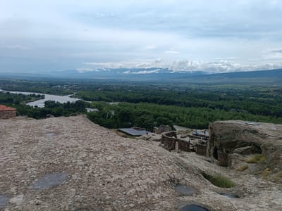 The image depicts a panoramic view of the ancient cave city of Uplistsikhe, Georgia, situated on a rocky outcrop overlooking a river and a lush green valley. In the foreground, large, weathered rocks with scattered puddles dominate the scene. Towards the midground, stone ruins of ancient structures are visible, with a few individuals exploring the site. Beyond the ruins, a dense forest stretches across the landscape, bordering a wide, light-colored river. In the distance, a town or village is nestled amongst the trees, leading to rolling hills and a mountain range under a cloudy sky. The overall impression is one of historical significance and natural beauty.