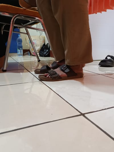 The image shows a pair of brown Birkenstock-style sandals on a person's feet, standing on a tiled floor. The person is wearing loose-fitting tan pants. In the background, a blue plastic water bottle is visible next to a chair with a metal frame. A black plastic bag and a patterned shopping bag are also in the background. To the right of the person's feet, a single black sandal is visible on the floor. The scene appears to be indoors, possibly in a home or public space.