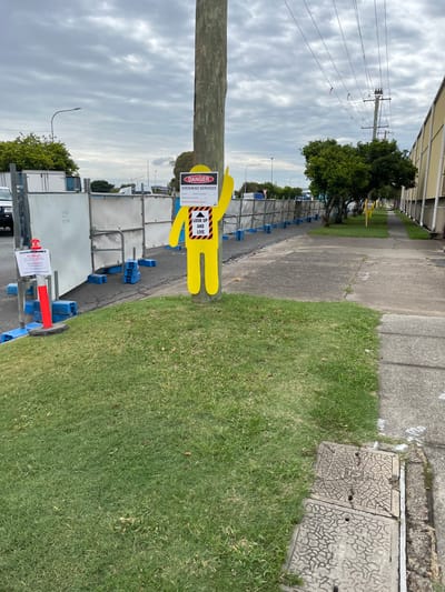 This image captures a street scene in Hamilton, Australia, primarily focusing on a safety installation related to overhead services. In the foreground, a tall, sturdy wooden utility pole stands prominently on the left. Attached to this pole is a bright yellow, human-shaped safety cutout, commonly known as a "Wobbly Man," positioned to face observers.

Two rectangular safety signs are affixed to the yellow figure. The upper sign, bordered in red and black, clearly displays "DANGER OVERHEAD SERVICES" in large letters. It further warns of "DANGER OF ELECTROCUTION," instructs to "KEEP CLEAR," and prohibits "NO LADDERS OR SCAFFOLDING OR MOBILE PLANT UNDER THIS LINE," with a partially visible line stating "Contact Ausgrid for further information." The lower sign, black with yellow text, features a distinctive downward-pointing arrow graphic above the bold directive "LOOK UP AND LIVE."

Adjacent to the utility pole, a concrete path or sidewalk extends into the distance, flanked by a strip of neatly maintained green grass. Further along the path, a series of white construction barriers or temporary fencing is visible, suggesting an active work zone or area requiring restricted access. In the midground and background, more utility poles with power lines stretch across the frame under an overcast sky. Some trees and what appear to be commercial or industrial buildings can be seen in the distance. On the far right, partially obscured by the grassy verge, are several grey, textured manhole covers embedded in the ground.

The photograph was taken during daytime, indicated by the overall lighting, though the sky is uniformly overcast, casting no distinct shadows. The combination of the utility pole, overhead lines, and safety signage strongly indicates an area with potential electrical hazards, where vigilance regarding overhead obstructions is crucial.
