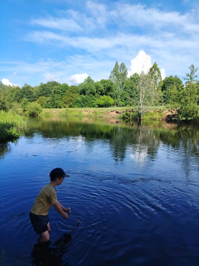 The image captures a boy standing in a dark blue body of water, likely a river or lake, on a sunny day. He is wearing a yellow t-shirt, dark shorts, and a baseball cap. The boy is holding a stick and appears to be playing in the water, disturbing the surface and creating ripples. The water is reflecting the blue sky, which is dotted with fluffy white clouds.

Behind the boy, the water extends to a shoreline lined with lush green trees and vegetation. The trees form a dense forest that creates a natural backdrop to the scene. The sun is high in the sky, suggesting it is midday. The overall mood is one of serene outdoor activity and a connection with nature. The location appears to be rural, with no visible man-made structures or signs of urban life. There is no visible text in the image.