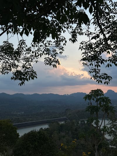 The image depicts an outdoor scene in Ban Ta Khun, Thailand, captured from an elevated viewpoint. A body of water is visible, partially obstructed by trees and foliage on the right side of the frame. A long concrete structure bisects the water, likely a dam or bridge. In the background, there are rolling hills covered in dense vegetation. The sky overhead is filled with clouds with hues of pink and blue. Silhouetted tree branches and leaves frame the upper portion of the image, primarily on the left. There are a few scattered lights visible in the distance along the hills.