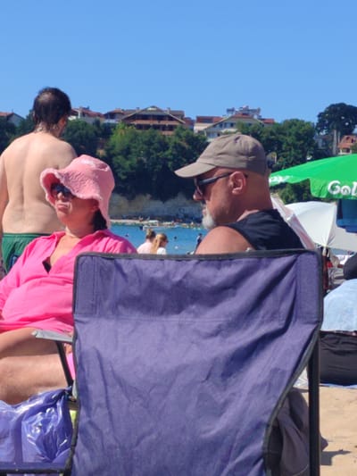 The image depicts a beach scene in Kandıra, Turkey. In the foreground, a woman wearing a pink sun hat and a pink top is seated in a purple camping chair. She is looking to her left with a slightly displeased expression. Behind her, partially visible, is a man wearing a cap and sunglasses, also seated in a chair. He is looking to his left. In the background, other beachgoers are visible, along with buildings and trees. A green umbrella with the partial text "Gol" is also present. The sky is clear and blue. The environment suggests a sunny day at the beach.