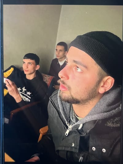 In Stepantsminda, Georgia, three young men are gathered indoors. The man in the foreground, wearing a black beanie and a black jacket over a grey hoodie, is looking to his right. To his left, a man in a black hoodie with white stars printed on it is seated and holding what appears to be a phone. Behind them, a third man in a dark jacket is also present. The room has plain walls and is sparsely furnished, with a patterned rug visible behind the seated man.