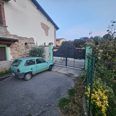 An exterior, low-angle, eye-level, wide shot captures a mint green Fiat Panda parked on an asphalt driveway in front of a large, ornate black metal gate. The gate has decorative scrollwork at the top and appears to be a driveway entrance. Above the gate, the letters "M L" are visible. To the left of the gate is a tall, slender, pale green pillar with a number "30" inscribed on it. Behind the pillar, the facade of a white building is visible, with sections of exposed brick and stone wall where plaster has chipped away. A brown wooden door with a light green frame is set into the left side of the building. To the right of the gate, a green wire fence encloses a patch of overgrown greenery, including a bush with bright yellow flowers. Beyond the gate and fence, trees and other buildings with terracotta roofs are visible against a pale blue sky. The overall scene suggests a rural or semi-rural setting in Calcinate, Italy, likely a residential property.