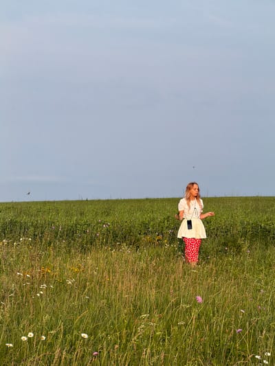 A young woman stands in a field of tall green grass and wildflowers under a pale blue sky. She is wearing a white short-sleeved shirt, a white skirt, and red patterned pants. A black smartphone is attached to a lanyard around her neck. The field appears to be in a rural area, possibly near Kutlu-Bukash, Russia, given the sparse vegetation and the open sky. A bird is visible in flight in the upper left quadrant of the image. The overall lighting suggests it is late afternoon or early evening.