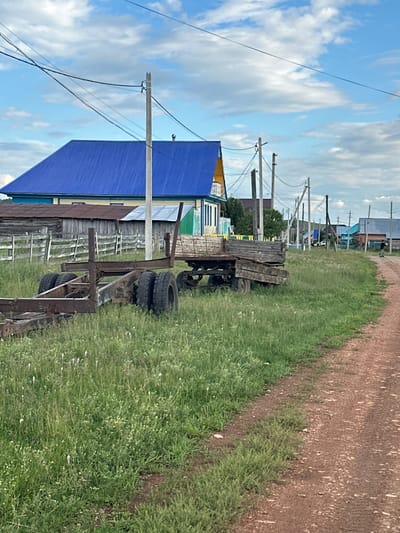The image depicts a rural scene in Urazovo, Russia, with a focus on a dirt road curving through a grassy area. In the foreground, two old, rusty trailers are parked amidst tall green grass dotted with small white and yellow wildflowers. The trailer on the left is a bare frame with four tires, while the trailer on the right is a wooden-bed structure.

Beyond the trailers, a wooden fence encloses an area with a weathered wooden building. Further in the background, a house with a bright blue metal roof stands out, adjacent to a yellow-sided structure. A series of utility poles with overhead wires run across the scene, with the sky above a mixture of blue and scattered white clouds.

The dirt road is reddish-brown and appears well-trodden. In the distance, along the road, several houses with colorful roofs are visible, suggesting a small village or settlement. A single person, dressed in dark clothing, is walking away from the camera on the dirt road in the middle ground, towards the right of the frame. The overall environment is one of a rural, possibly somewhat neglected, agricultural area.