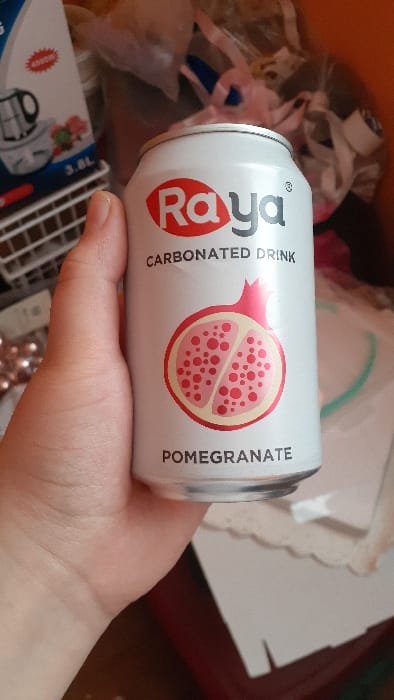 A hand holds a can of Raya pomegranate carbonated drink. The can is white with red logo and a pomegranate graphic. The background is cluttered with various objects including a 3.8L container, wrapping paper, and a small rack. The overall setting suggests a crafting or gift-wrapping area.
