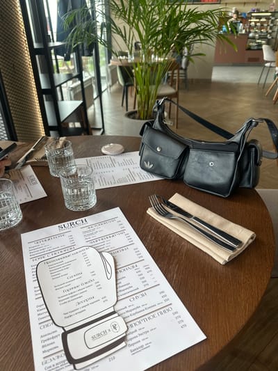 The image depicts a dining table within a cafe or restaurant in Усады, Russia. A round wooden table dominates the foreground, upon which two clear, ridged glasses filled with water are placed. A white menu, printed in Russian with the "SURCH COFFEE & KITCHEN" logo, is spread across the table, partially obscured by a black leather shoulder bag with multiple exterior pockets. One pocket of the bag displays a silver Adidas logo. To the right of the menu and bag, a set of silver cutlery (fork and knife) rests on a folded beige napkin. A small, smooth white object is visible near the top edge of the menu. A hand, likely holding a mobile device, is partially visible on the far left.
The background reveals a brightly lit cafe interior with large windows along the far wall, through which greenery is visible. Various tables and chairs are sparsely arranged, suggesting a spacious layout. A large potted plant with numerous long green fronds stands prominently in the mid-right. In the blurred distance, a counter area with display cases and faint human figures is discernible. On the far left, a black metal shelving unit and a textured screen are visible, with a dark jacket hanging on the shelf. The floor appears to be light-colored concrete or tile.