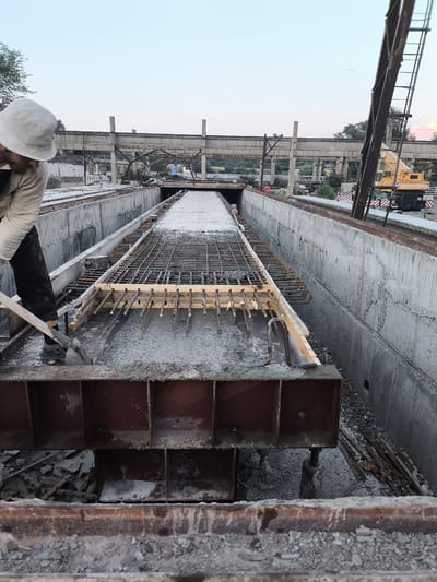 A construction worker, wearing a white bucket hat and dark pants, is using a shovel to spread wet concrete in a trench. The trench is lined with concrete walls and filled with rebar, indicating an ongoing construction project. The scene appears to be outdoors, with a bright sky suggesting it is either early morning or late afternoon. In the background, industrial structures, including concrete pillars and beams, are visible, along with a yellow crane. The environment suggests an industrial or infrastructure construction site. The worker's posture and the act of spreading concrete point to the task of laying a foundation or a concrete surface. The overall mood is one of diligent work. There is no visible text in the image.