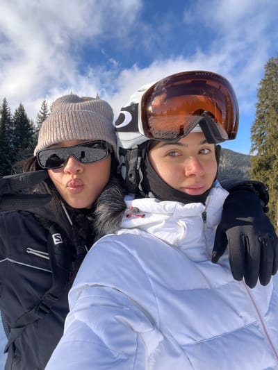 Two individuals, positioned in a close-up, chest-up outdoor photograph, are situated in a snowy, mountainous environment in Saanen, Switzerland. The person on the left wears a light brown knitted beanie, black wraparound sunglasses, and a black winter jacket featuring a dark fur-lined hood. Their left hand, covered by a black glove, displays two extended fingers in a V-shape, and their lips are pursed. A white "Salomon" logo is visible on the left side of their jacket. The person on the right wears a white ski helmet adorned with a black and white "Oakley" logo, and large amber-tinted ski goggles that reflect the surrounding snowy landscape and blue sky. A black neck gaiter covers their lower face. This individual is dressed in a white padded winter jacket with visible red zipper pull tabs and black gloves, with their right arm positioned behind the left individual. Both subjects are looking towards the camera. The background consists of a clear blue sky with scattered white clouds above a line of snow-dusted evergreen trees, indicating a winter landscape.