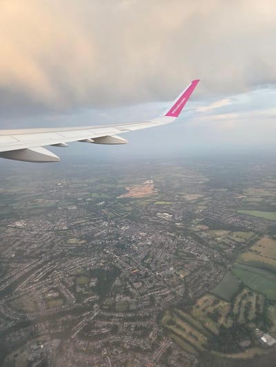 The image shows an aerial view from a Wizz Air plane, with the plane's wing visible on the left side of the frame. The ground below consists of a mixed landscape of urban and rural areas. A dense network of roads and buildings suggests a residential area. Several green spaces, including what appears to be a golf course, are also visible. Based on the pattern of houses and green spaces, the city is most likely St Albans, United Kingdom.