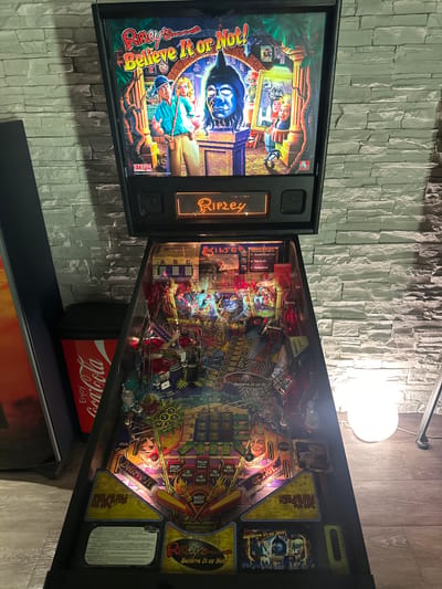 An indoor scene in Winterthur, Switzerland, captures a brightly lit "Ripley's Believe It or Not!" pinball machine from a slightly elevated, side perspective. The pinball machine is prominently displayed against a textured wall made of light-colored, stacked stone or brick-like panels.
The pinball machine's backglass features colorful artwork depicting characters and scenes typical of Ripley's attractions, with "Ripley's Believe It or Not!" and "STERN" logos clearly visible. The playfield, rich with intricate illustrations and numerous targets, lights, and ramps, glows with vibrant colors. Artwork on the playfield includes jungle/adventure themes, various characters, and a skull figure. Several game features and scores are labeled, such as "SPECIAL," "EXTRA BALL," "BIG ROAR," "SUPER JACKPOT," and various "MILLION" point markers. Text like "SHOOT AGAIN," "COLLECTED STONE," "SKILL," and "PRESS AND HOLD BUTTON FOR INFO" are also visible. A list of possible events like "A. Scramble Temple Code," "B. Wake Up Idol," and "C. Amazing Event!" is displayed.
Below the playfield, a red Coca-Cola box or cooler, with "Enjoy Coca-Cola" printed upside down, rests on a dark shelf or cabinet. A spherical white lamp, casting a warm glow, is visible in the upper right portion of the image, contributing to the overall illumination of the scene. The pinball machine appears to be on, suggesting it is either in attract mode or ready for play, though no people are present.