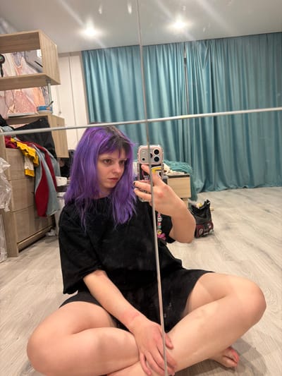 A person with purple hair is sitting cross-legged on a light wooden floor, taking a selfie in a mirror. The individual wears a black t-shirt and black shorts. The background includes a blue curtain, a light brown wooden table, and a gray table with assorted items. A pair of shoes are visible on the floor. The person holds a cellphone in their right hand, capturing their reflection.