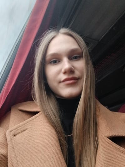 A young woman with long blonde hair and light skin is shown in a selfie-style photograph. She wears a black turtleneck sweater and a tan overcoat. The background suggests she is inside a vehicle; a maroon surface is visible behind her head, and a window is to her right. The photograph is oriented at a slight angle. No specific details are discernible to confirm the location as Chernovskoe, Russia. The photo solely shows a young woman inside a vehicle.