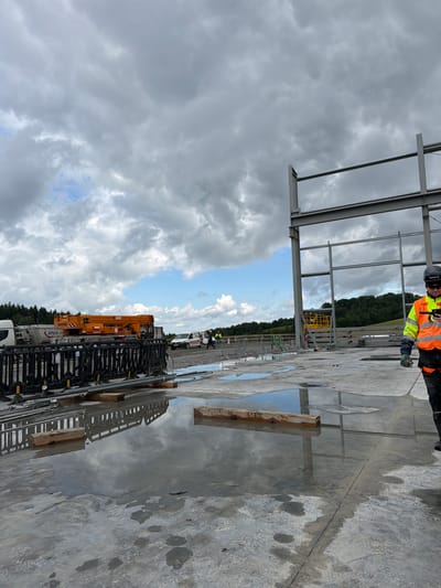 The image depicts a construction site under a cloudy sky. In the foreground, a construction worker wearing a high-visibility orange vest and a hard hat is walking towards the right side of the frame, holding a mobile phone. The ground is wet and reflects the sky and surrounding structures. To the left, a line of temporary barriers is visible, with a large orange crane and several vehicles behind them. In the background, a partially constructed metal building frame is apparent, along with trees and a gently sloping landscape. The scene suggests an active construction project.
