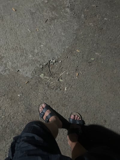 The image depicts a patch of concrete ground with a rough texture. Debris, including small rocks and dried leaves, is scattered across the surface. A pair of feet wearing black sandals are visible on the right side of the frame. The lower portion of a person's legs and black shorts are also visible, indicating the person is standing. A shadow partially obscures the scene on the upper right side. There is no information given in the picture to confirm the location in Binong, Indonesia.