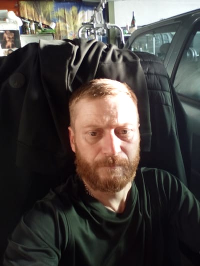A man with reddish-brown hair and a beard, wearing a dark green t-shirt and a dark jacket, is pictured from the chest up. He is looking towards the camera. The background appears to be a garage or workshop, with various objects visible including shelves with items, a car, and a metallic structure. The lighting suggests it is indoors. This image is not located in the city of Rottweil, Germany.