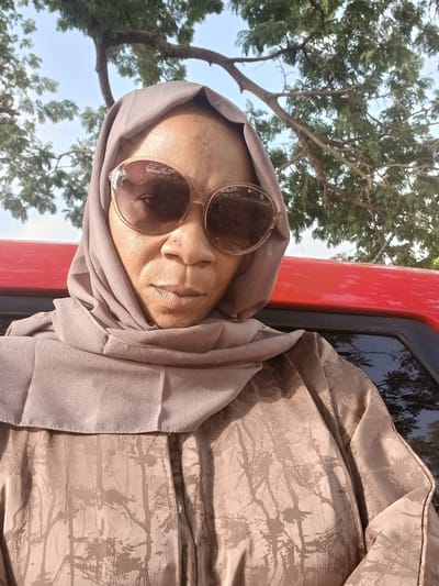 A woman with a light brown complexion wearing a light brown headscarf and brown sunglasses is posing for a photo. She is partially seen inside a red vehicle, perhaps a car, with a window visible next to her. Outside, a tree with green leaves is visible against a partly cloudy sky. The location is Rigachikun, Nigeria.