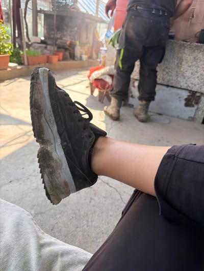 A medium-shot captures a person's leg and foot wearing a dirty black and grey sneaker. The person is dressed in black, possibly shorts or pants. A blurry background reveals a cluttered outdoor scene, potentially a greenhouse or garden in disarray with exposed beams, potted plants, and other indistinct objects. A second person is visible with their legs in black pants and work boots, along with a red bucket. This scene is presumed to be near or in Damianitsa, Bulgaria, given the provided information.