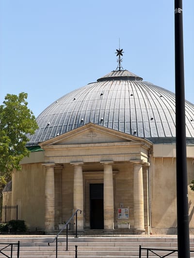 This image, taken in Courbevoie, France, features a grand architectural edifice, likely a church or public building, with a prominent domed roof. The dome is constructed of metal, appearing to be silver or gray, and is segmented into numerous panels. At the apex of the dome, a decorative metal spire with a star-like finial is visible against a clear, bright blue sky.

The building's facade is made of stone, with large columns supporting an upper level. A set of wide stone stairs leads up to an entrance with a dark door, flanked by more stone columns. A metal railing runs along the side of the stairs, providing a visual guide.

Green foliage from trees is present at the base of the building and also peeks out from behind the dome, suggesting a pleasant outdoor setting. The sunlight casts shadows, indicating it is likely daytime, possibly late morning or afternoon, given the angle of the light. The overall atmosphere is one of grandeur and tranquility, with no people visible. There are no discernible emotions, and the weather appears clear and fair.

A poster or notice board is affixed to the wall near the entrance, displaying various pieces of colorful text and images, but the content is not legible. A black pipe or beam runs horizontally across the top of the image, partially obscuring the view of the sky and the top of the dome, possibly part of scaffolding or an overhead structure.