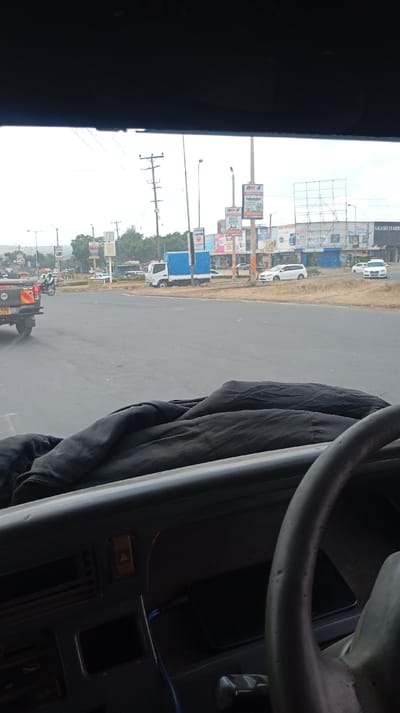 An interior view from a vehicle reveals a road scene. A white truck with a blue tarp and a motorcycle with police markings are visible on the left. Billboards and power lines punctuate the horizon. The road is dry. A white car is visible on the right. Part of the dashboard, a dark covering, and the steering wheel are in the foreground. A phone is visible on the dashboard.