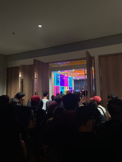 The image depicts a gathering of people in what appears to be an entertainment venue in Salembaran, Indonesia. The room features a colorful, illuminated floor composed of squares in shades of blue, pink, orange, and yellow. People are standing in a line or gathered in the room, but the view is partially obstructed by individuals in the foreground. The lighting is dim, with a strong contrast between the illuminated floor and the darker areas of the room and people. Wooden architectural elements, possibly door frames or wall panels, are visible around the opening of the space.