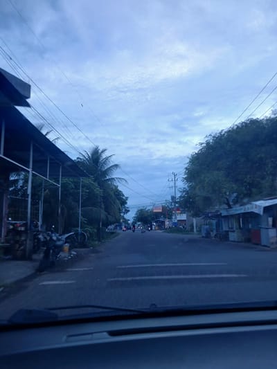 This is a slightly out-of-focus, horizontal, medium-shot, point-of-view photograph taken from inside a vehicle, looking forward at a paved road in Geulanggang Teungoh, Indonesia. The sky is overcast with pale blue and white clouds.

On the left side of the road, there is a long, open-sided structure with a metal frame and a corrugated roof. Underneath this structure, several motorcycles are parked, some with visible helmets and another with a yellow bucket nearby. Palm trees and other greenery are behind this structure, creating a dense natural barrier. Power lines run diagonally across the sky, supported by utility poles visible in the distance.

The road itself is asphalt with white dashed lane markings. Several motorcycles and a few other vehicles are traveling on the road, heading into the distance. On the right side of the road, there are small, simple buildings or stalls with awnings and some appear to be shops. One stall has a red and blue facade. A sign with the "Alfamart" logo is visible on a building further down the road on the left.

The foreground of the image shows the dashboard and windshield of the vehicle, which are dark and partially obstruct the view. The overall lighting suggests it is either early morning or late afternoon, given the dim natural light and cloudy conditions.