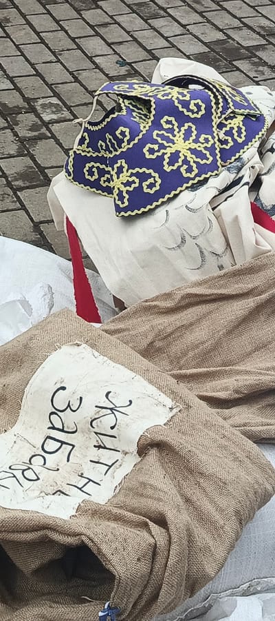 The image shows a close-up view of several burlap sacks and some fabric items piled on a cobblestone surface. The sacks are a natural brown color and appear to be full, with one sack open at the top, revealing its contents.
On top of the sacks, there are several pieces of fabric. One is a cream-colored fabric with a pattern of dark, curved lines that resemble scales or waves. Another is a piece of bright red fabric. The most prominent fabric item is a purple cap or hat with intricate yellow embroidery, featuring a floral or star-like motif and a decorative trim. This cap is placed on top of some of the other fabrics.
In the foreground, a torn piece of white paper or fabric is attached to one of the burlap sacks. It has handwritten text in black ink. The visible text appears to be in Cyrillic script and reads "Живу я забере" or a similar combination of letters. The writing is in a somewhat stylized, cursive font.
The setting appears to be outdoors, suggested by the cobblestone ground which looks like a pathway or courtyard. The lighting is somewhat dim, indicating it might be overcast or late in the day, though it's difficult to determine definitively. There are no people visible in the image. The overall impression is of items that have been gathered or stored, possibly in a market or storage area. The presence of traditional-looking clothing or headwear (the purple cap) alongside everyday sacks might suggest a cultural or commercial context related to local crafts or goods.