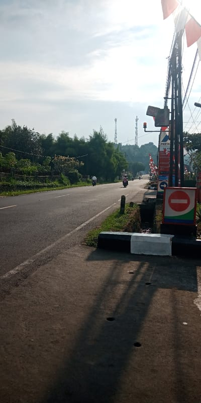 The image depicts an outdoor roadside scene in Cijedil, Indonesia, under a bright sky with the sun positioned high on the right, casting long shadows across the foreground.
The scene is bisected by an asphalt road, marked with a solid white line on the right shoulder and a broken white center line, extending into the distance with a gentle upward slope. On the left side of the road, dense green vegetation, including bamboo and other trees, is visible behind a low concrete wall or fence. Two motorcycles are actively traveling away from the camera on the road; one, appearing light-colored, is further in the distance, and another, darker, is closer. A third, more distant motorcycle is faintly visible.
The immediate foreground on the right features a paved shoulder with dark, elongated shadows. A black and white painted concrete barrier is present, adjacent to an open drainage ditch. Beyond the ditch, a complex utility pole structure rises, supporting numerous overhead wires, a small orange flashing light, and an electrical junction box. Attached to this pole assembly are several signs: a blue "PERTAMINA" sign listing "PERTAMAX TURBO," "PERTALITE," and "PERTAMINA DEX"; a blue circular directional sign with a white arrow pointing right; and a red circular "No Entry" sign with a white horizontal bar. Red and white striped banners or flags are partially visible on the far right, near the sun's glare. In the background, tree-covered hills are visible, topped by two distinct cellular or telecommunication towers.