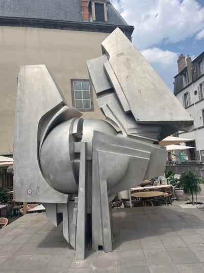 This is a photograph of a large, abstract metal sculpture located in a plaza in front of buildings in Clermont-Ferrand, France. The sculpture is composed of interlocking geometric shapes and a central sphere, all rendered in brushed metal. The plaza pavement is made of gray square tiles. In the background, there are buildings with visible architectural details such as windows and chimneys. Outdoor seating areas with tables and chairs are visible, some with umbrellas. A few potted plants are also present. The sky is partly cloudy. The sculpture is the primary subject of the image, dominating the foreground. No people are clearly identifiable in the immediate vicinity of the sculpture.