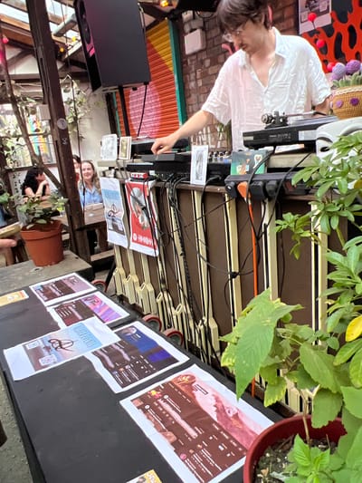 The image captures a vibrant scene, likely at an outdoor event or gathering, with a strong emphasis on music and community. In the foreground, a person is partially visible, appearing to interact with DJ equipment, suggesting a DJ set or music performance is underway. They are wearing a white shirt and have dark hair. Scattered across a table are various printed materials, including what appear to be social media profiles or event flyers. One flyer prominently features the text "ALINEA EVENTS" along with a stylized logo. Other visible text includes "Spotify" on a red flyer, and what looks like profile information with user names, follower counts, and post snippets on several other printed pages. The background reveals a lively, perhaps bohemian or urban setting with colorful corrugated metal sheeting on a wall, brickwork, and abundant greenery from potted plants. The overall atmosphere seems energetic and social, with the presence of multiple people, though some are only partially visible or blurred. The lighting suggests it could be daytime or early evening. The context provided places this scene in Liverpool, United Kingdom.