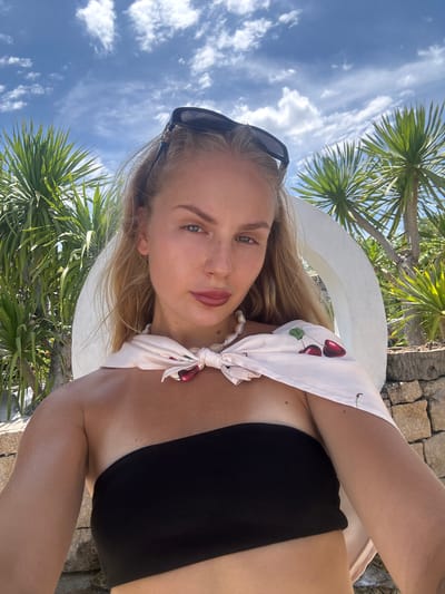 A young woman is taking a selfie outdoors in a tropical location. She is wearing a black bikini top and a light-colored scarf tied around her neck with a cherry print. She has on sunglasses pushed up on her head. The background shows palm trees and a bright blue sky with scattered white clouds, suggesting it is daytime with pleasant weather. The setting appears to be a resort or a sunny outdoor area, possibly near water given the style of dress and vegetation. Her expression is relaxed and natural, typical of a casual selfie. There is no visible text in the image.