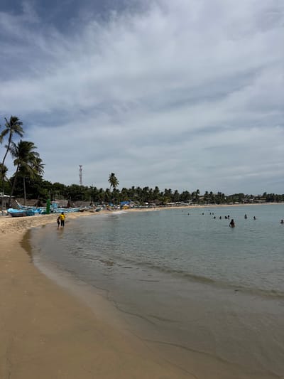 The image captures a scene at a beach in Arugam Bay, Sri Lanka, under a partly cloudy sky. The foreground features a sandy beach with gentle waves lapping at the shore. Two individuals, one appearing to be an adult in a yellow shirt and a child, are wading in the shallow water. Further down the beach, a line of blue and white fishing boats are pulled ashore. Behind the boats, palm trees and low-lying buildings line the coast, suggesting a village or resort area. In the middle ground, several people are swimming and relaxing in the ocean. The background shows a densely vegetated coastline with more palm trees and distant structures. A telecommunications tower is visible above the tree line, along with a communication antenna. The overall atmosphere is one of a tranquil coastal setting with recreational activities taking place.