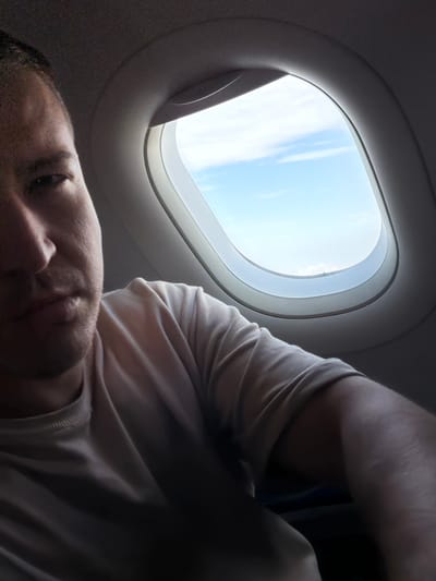 A man is looking into an airplane window. The window is oval-shaped with a white frame. Outside the window, there is a clear blue sky with some wispy clouds. The man is wearing a light-colored t-shirt. The image is taken from a low angle, showing the man's face and upper body. There is no identifiable indication of location in the city of Mohan, India.